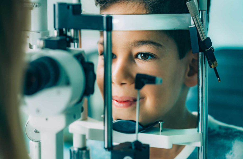 A close up of a young boy smiling with his head on a chin rest while an optometrist performs a slit lamp test