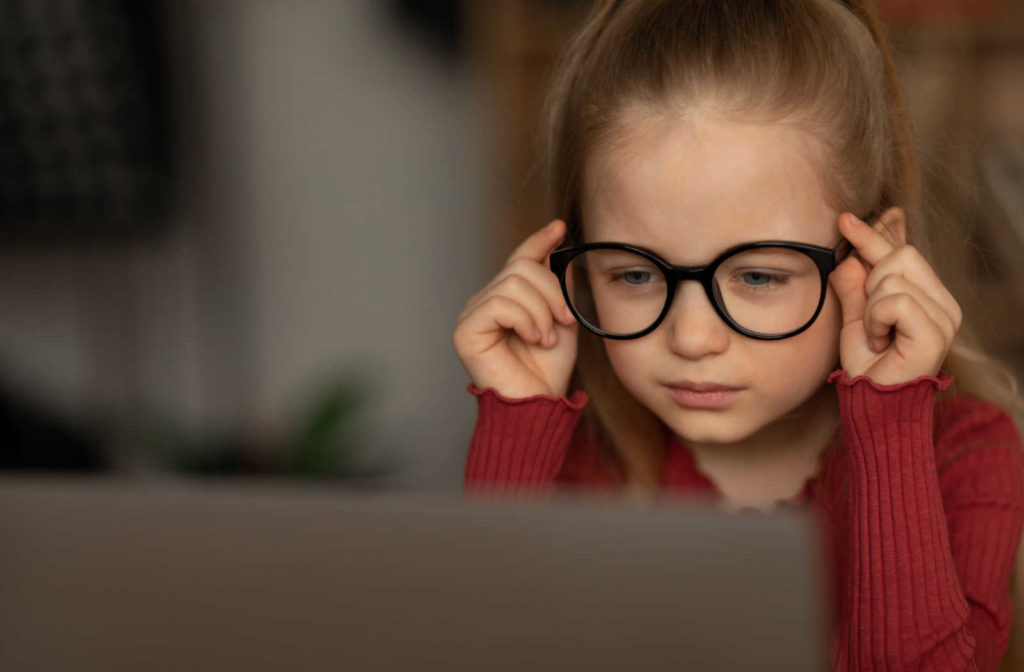 A female preschooler in a red long-sleeve shirt with her eyeglasses is sitting in front of her laptop computer with a gesture on her face of having a hard time reading.