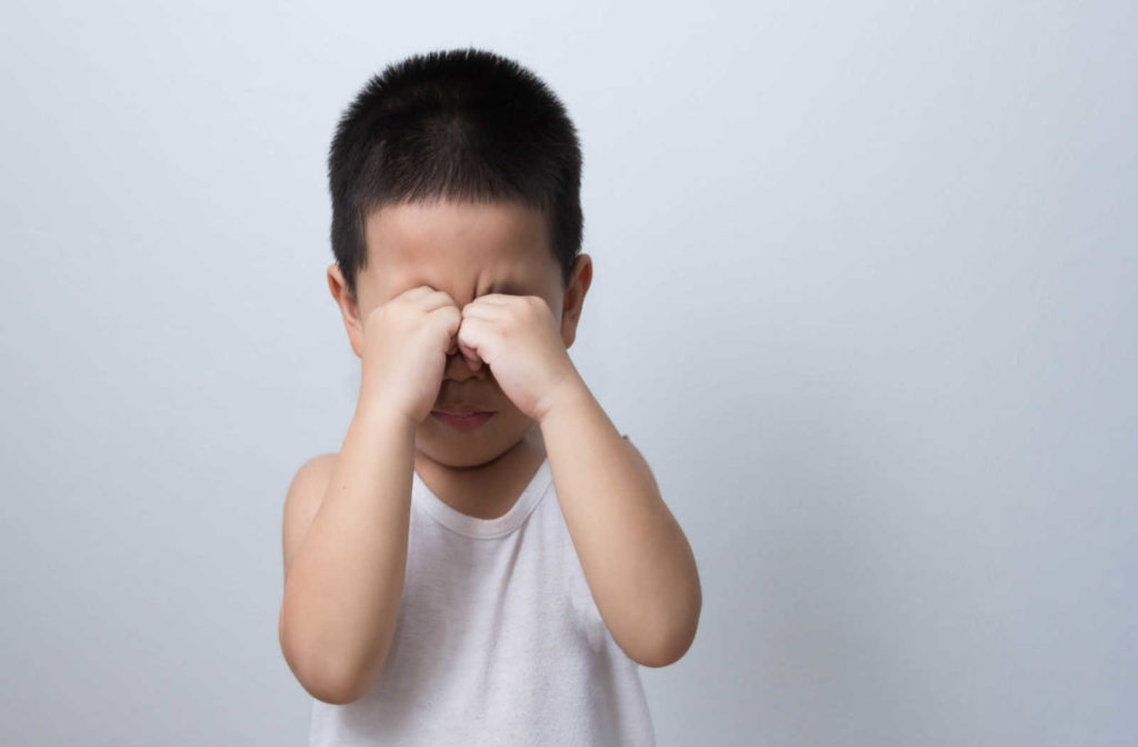 A young boy in a white shirt rubbing his eyes with his fists