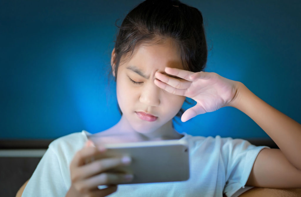 A young girl is rubbing her irritated left eye due to long exposure to blue light while using her cell phone.