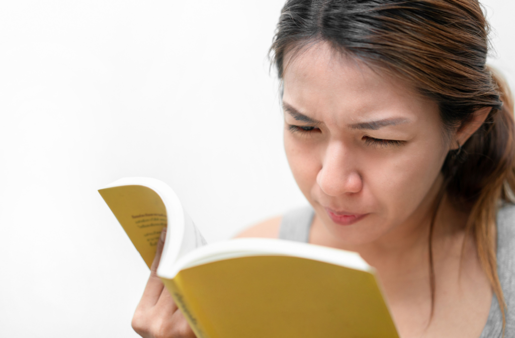 A young woman squinting her eyes to see more clearly while holding a book on her right hand.