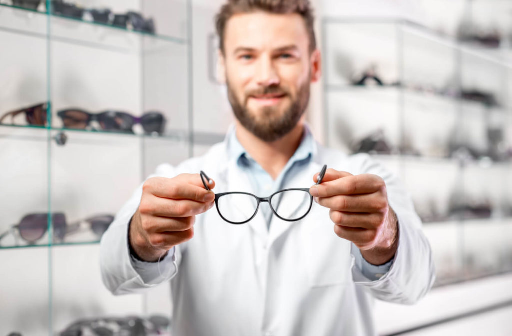 An optometrist holding a pair of prescription glasses as he smiles and looks directly at the camera.