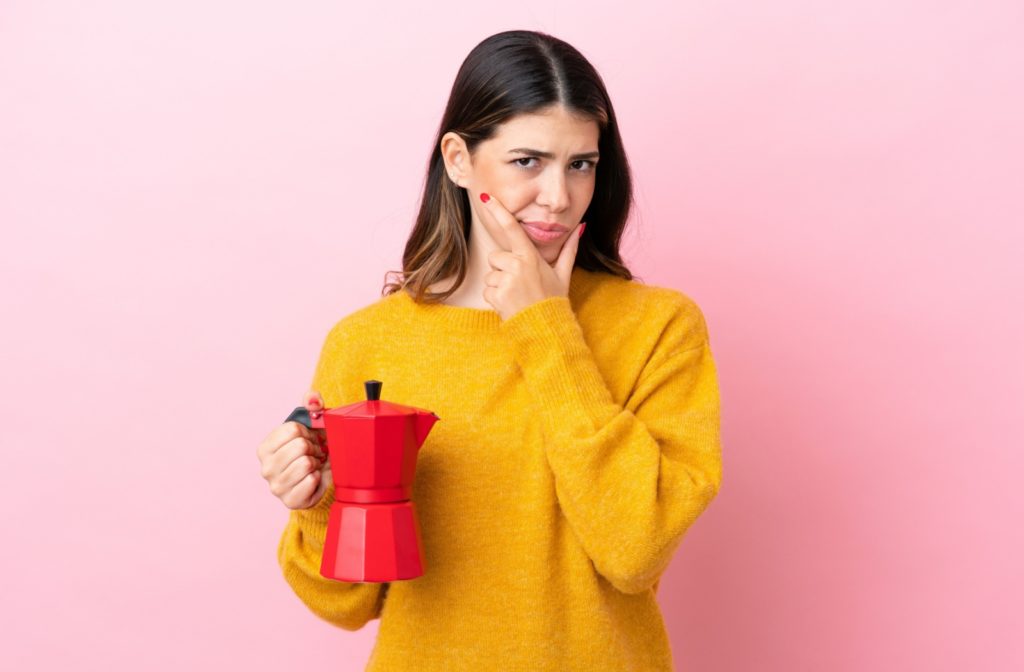 A woman in front of a pink background holding a coffeemaker trying to decide if she wants more coffee.