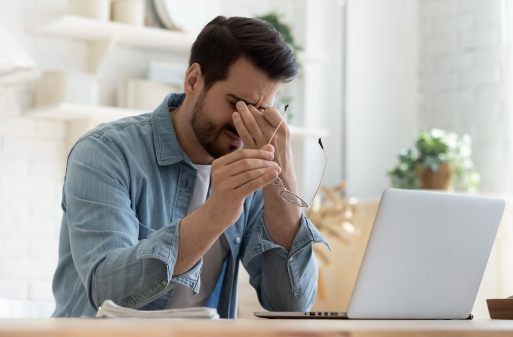 A young man taking off his glasses and rubbing his eyes while working due to discomfort from sinusitis.