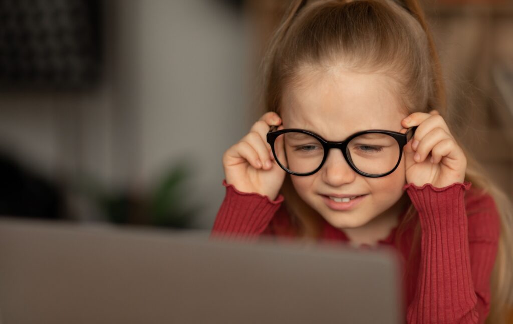 A young child wearing thick-framed black glasses squints and looks closely at the computer screen in front of them.