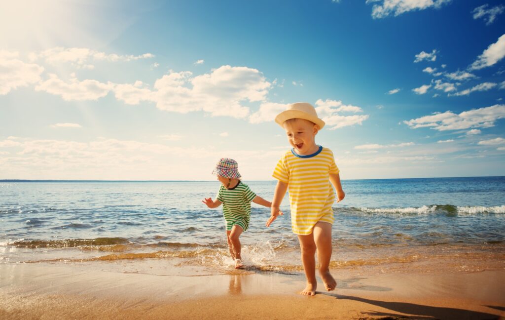 Two children wearing hats and large t-shirts run and play at the edge of the water at a beautiful beach.