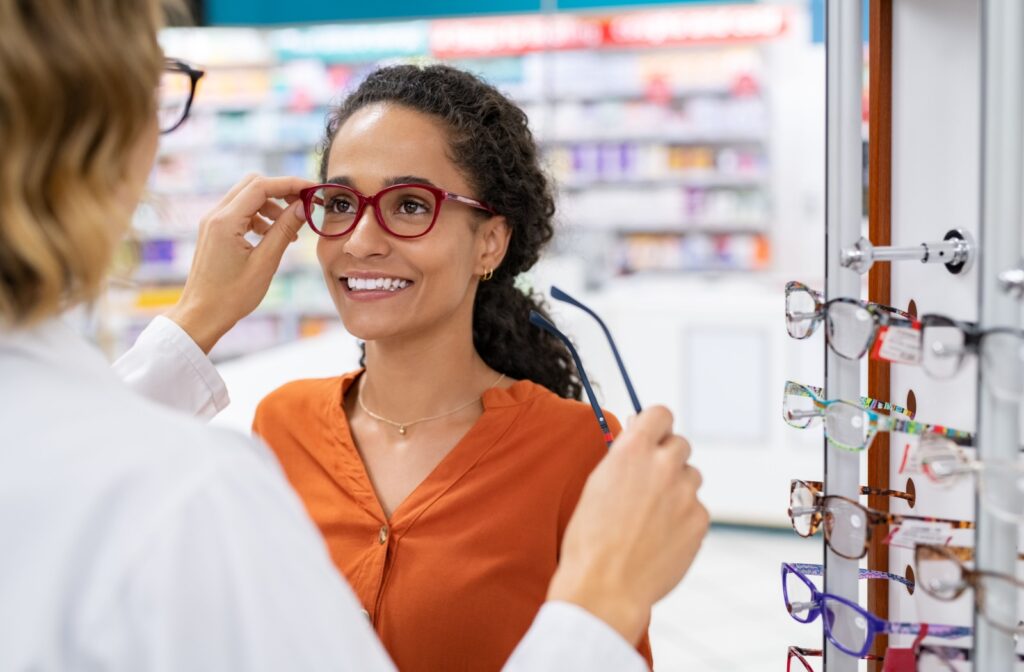A person tries on red eyeglass frames with help from an eye care professional, standing beside a display of glasses in an eyewear store.