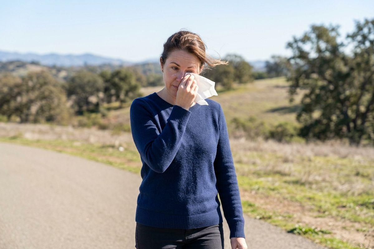 Person in blue sweater wiping eye with a tissue while standing on a rural path with hills and trees behind.